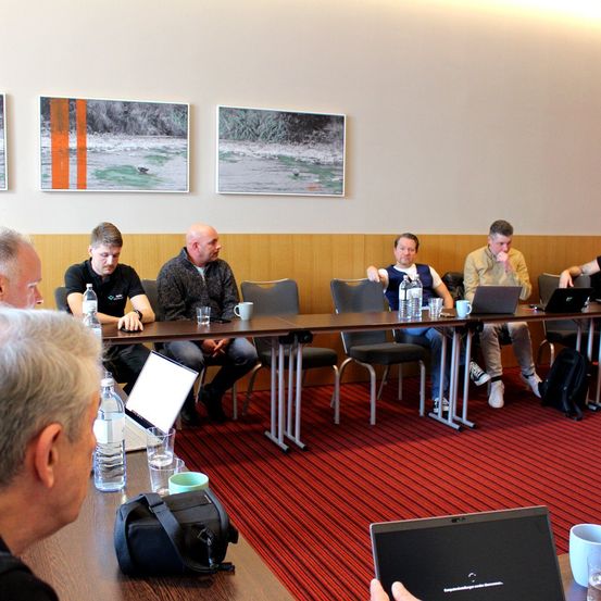 Eine Gruppe von Männern sitzt um einen Tisch mit Laptops, Wasserflaschen und Tassen. Sie scheinen sich in einem Konferenzraum mit rotem Teppich und drei Gemälden an der Wand zu befinden.