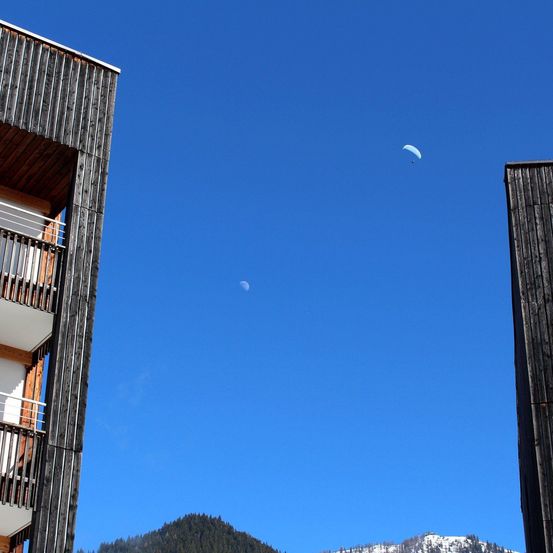Ein Gebäude mit Balkonen steht unter einem klaren blauen Himmel. Ein Gleitschirmflieger schwebt in der Nähe eines entfernten Mondes. Im Hintergrund sind schneebedeckte Berge zu sehen.