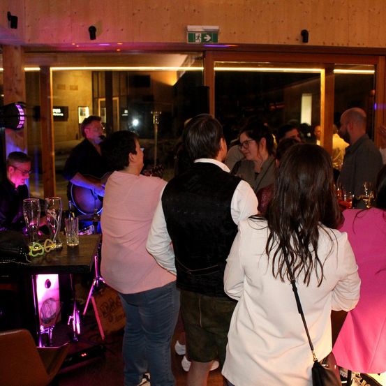 Eine Gruppe von Menschen ist in einem Club, ein Mann spielt Gitarre. Der Raum ist mit lila Lichtern beleuchtet und es stehen Gläser auf einem Tisch.