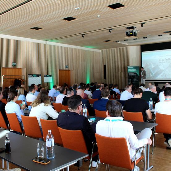 Zuhörer sitzen in Reihen vor einer Projektionsleinwand. Ein Mann steht an einem Podium vor einem grünen Hintergrund. Der Raum hat Holzwände und Tische.
