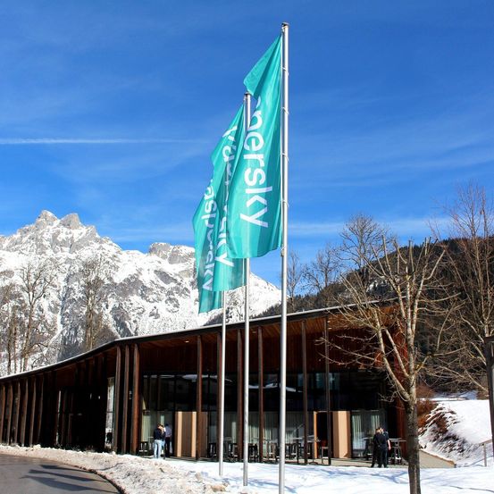 Ein modernes Gebäude mit schneebedecktem Dach steht vor einer Bergkette. Drei Fahnen, darunter eine der Firma 'Persky', sind auf Stangen gehisst. Es gibt Menschen vor dem Gebäude.