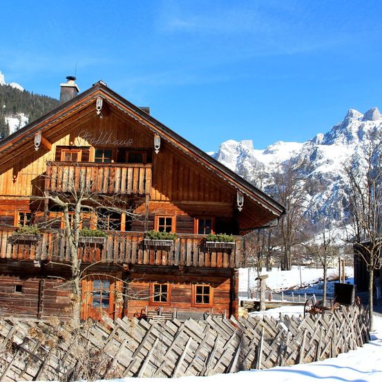 Ein Holzchalet mit Balkonen und Schornsteinen, eingebettet in verschneite Berge, umgeben von einem Holzzaun in einer verschneiten Landschaft.