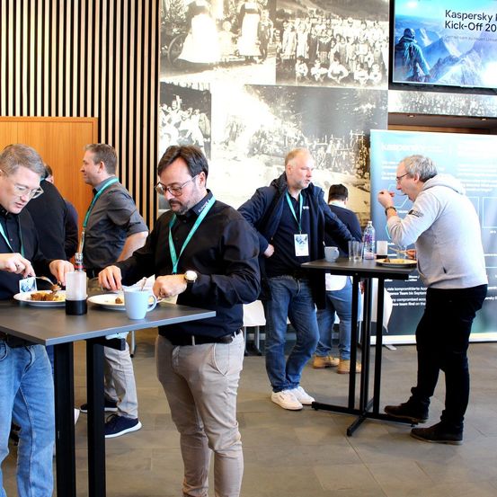 Men in Anzug und Krawatte versammeln sich auf einer Konferenz, einige sitzend und essen, andere stehend. Im Hintergrund befindet sich ein großes Foto mit dem Schriftzug 'Kaspersky Kick-Off 20'.