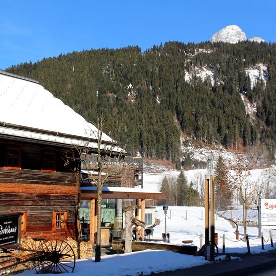 Ein holzverkleidetes Gebäude mit schneebedecktem Dach steht in der Nähe eines schneebedeckten Berges mit Bäumen. Links befindet sich ein Schild, und vor dem Gebäude steht ein Wagenrad.