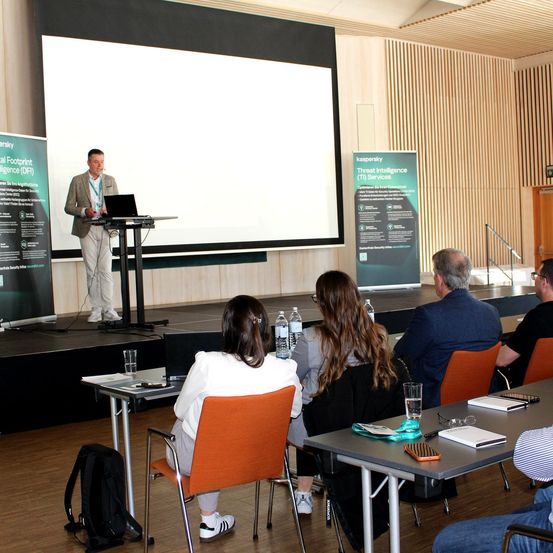Ein Mann hält eine Präsentation in einem Konferenzsaal mit einem Laptop auf einem Podium. Mehrere Personen sitzen vor ihm und hören aufmerksam zu. Es gibt Wasserflaschen, Notizbücher und andere Gegenstände auf dem Tisch.