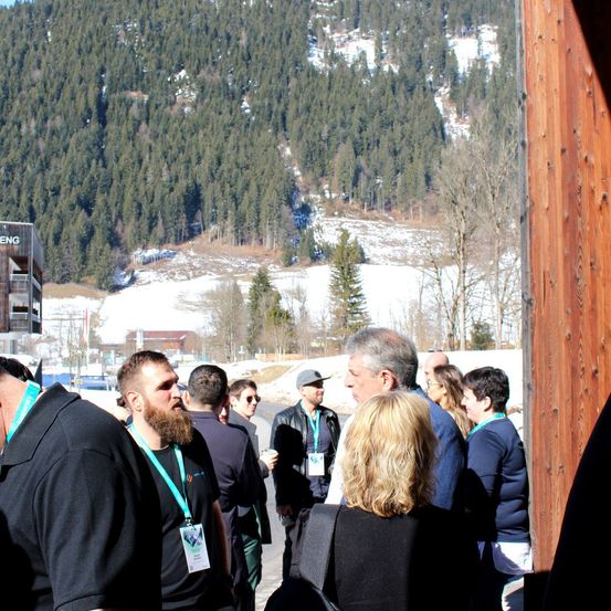 Eine Gruppe von Menschen mit Namensschildern steht im Freien in einer Schlange mit einem verschneiten Berg im Hintergrund.