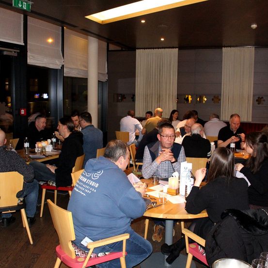 Eine Gruppe von Menschen sitzt an Tischen in einem Restaurant. Sie führen Gespräche und einige benutzen ihre Telefone. Der Raum ist schwach beleuchtet mit weißen Vorhängen. Flaschen und Gläser stehen auf den Tischen.
