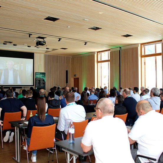 Ein Konferenzraum mit einer großen Leinwand, auf der ein Mann zu sehen ist. Die Teilnehmer sitzen in Reihen und hören aufmerksam zu, mit einem Projektor und einem Schild an der Wand.
