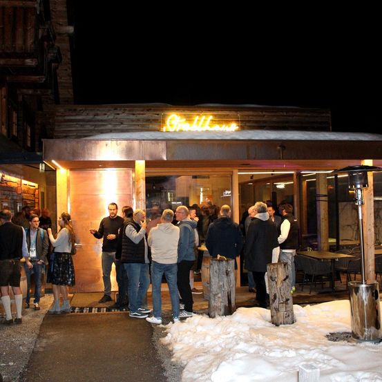Mehrere Menschen stehen nachts vor einem Restaurant, das von gelbem Licht beleuchtet wird. Der Name des Restaurants lautet Grillhaus. Sie sind warm gekleidet. Einige Leute reden und lachen. Es gibt Tische und Stühle im Restaurant. Auf der rechten Seite steht ein großer Holzpfosten.
