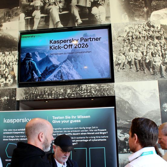 Ein großer Monitor zeigt 'Kaspersky Partner Kick-Off 2026'. Darunter schaut eine Gruppe von Männern auf einen Bildschirm mit Text zum Testen ihres Wissens. Ein Mann in einem Hut schaut sie an.