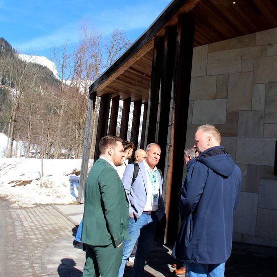 Eine Gruppe von Männern in Anzügen steht vor einem Gebäude mit Holzbalken und Steinmauern, mit verschneiten Bergen im Hintergrund.