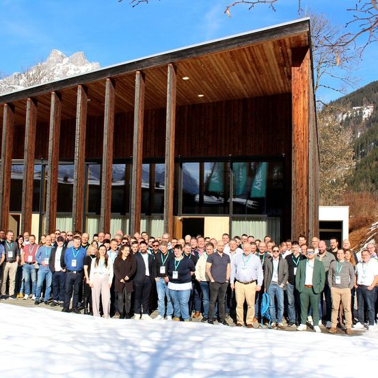 Eine große Gruppe von Menschen mit Ausweisen steht vor einem Holzgebäude mit Glasfenstern. Sie stehen auf dem Schnee vor einem Gebäude mit Bergen im Hintergrund.