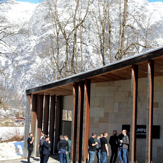 Eine Gruppe von Menschen steht vor einem modernen Gebäude mit einem geneigten Dach, in der Nähe eines schneebedeckten Berges. Die Personen unterhalten sich.