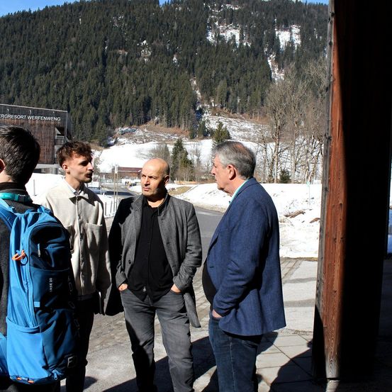 Vier Männer stehen im Freien mit einem Berg im Hintergrund. Der Mann in blauem und grauem Anzug führt ein Gespräch, während ein anderer Mann mit einem Rucksack zuhört.