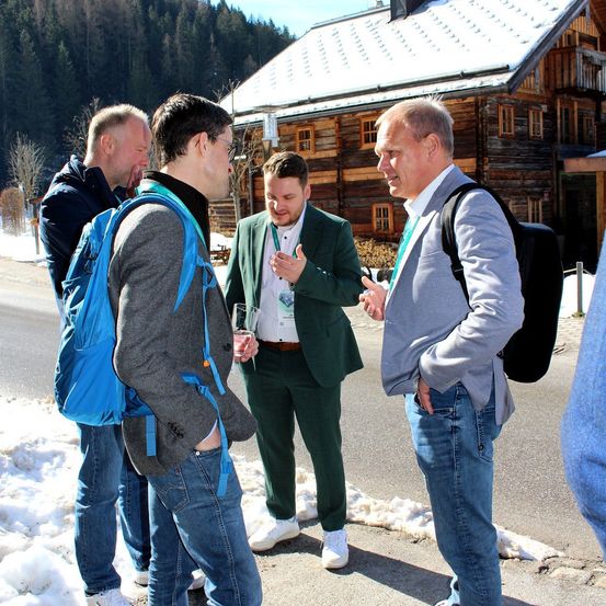 Vier Männer stehen im Schnee draußen, tragen Mäntel und Jeans. Sie scheinen ein Gespräch zu führen. Hinter ihnen steht ein Holzhaus.