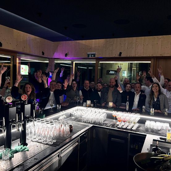 Eine Gruppe von Menschen feiert in einer Bar, hebt Gläser und posiert für ein Foto. Die Bar ist gut beleuchtet mit modernem Design, mit einer großen Theke mit Gläsern und Flaschen.