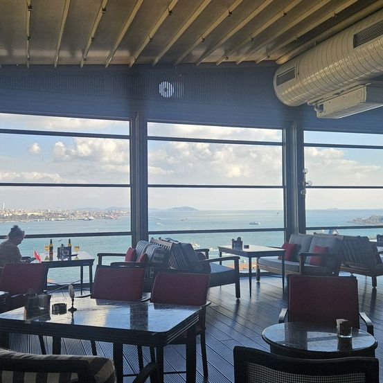 Ein gehobenes Restaurant mit großen Fenstern, mit Blick auf das Meer und die Stadt. Tische und Stühle sind aufgestellt, eine Person sitzt an einem Tisch. Flaschen, Gläser und verschiedene Gegenstände sind auf den Tischen platziert. Der Raum ist von Sonnenlicht durchflutet.