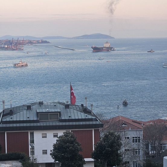 Luftaufnahme einer Stadt mit mehreren Booten und einem großen Schiff, das auf dem Meer fährt. Eine türkische Flagge ist auf einem Gebäude gehisst.