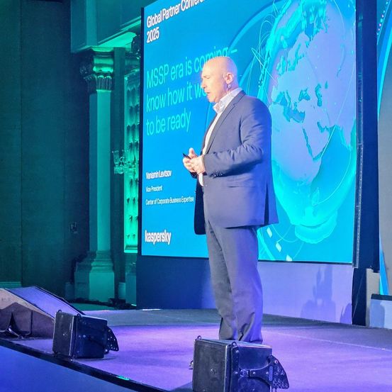 Ein Mann steht auf der Bühne und hält eine Präsentation. Hinter ihm ist eine große Leinwand, auf der der Text 'Global Partner Conference 2025' zu sehen ist. Der Mann ist in einem formellen Anzug gekleidet.