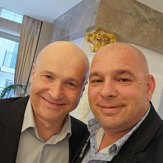 Zwei Männer stehen eng beieinander und lächeln in die Kamera. Einer trägt ein graues Hemd mit einem weißen Kragen, der andere trägt einen schwarzen Anzug mit einem weißen Innenshirt.