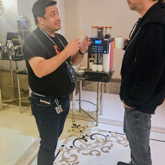 Zwei Männer stehen in einer Hotellobby neben einer Kaffeemaschine, einer hält eine Kaffeetasse. Der Mann mit dem Ausweis spricht.