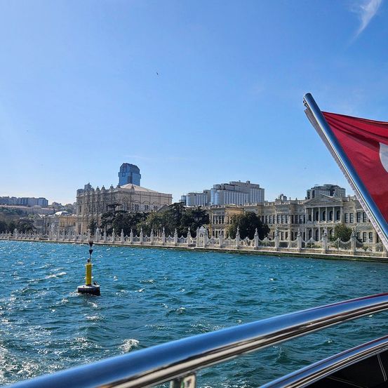 Ein Boot mit einer türkischen Flagge fährt an der Küste entlang mit Blick auf ein großes Gebäude. Das Wasser ist ruhig und es gibt einen Bojen im Wasser.