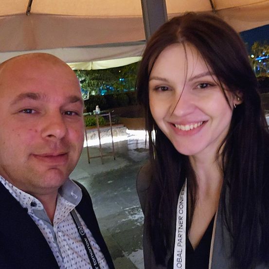 Ein Mann und eine Frau lächeln in die Kamera, stehen unter einem Zelt mit Lichtern und Grünpflanzen im Hintergrund. Die Frau trägt eine Umhängetasche, auf der 'Global Partner Conference' steht.