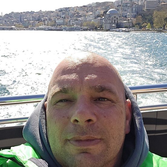Ein Mann in einer grünen Jacke und einer grauen Kapuzenpulli sitzt lächelnd auf einem Boot, mit einer Skyline im Hintergrund.
