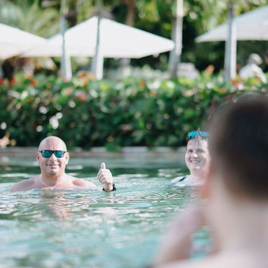 Drei Menschen schwimmen in einem Pool. Ein Mann gibt ein Daumen-Hoch-Zeichen. Die Frau neben ihm lächelt. Dahinter blickt eine verschwommene Person auf sie.