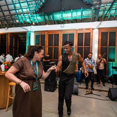 Ein Mann und eine Frau tanzen in einem hell beleuchteten Raum, mit einer Band im Hintergrund. Die Band spielt Gitarren und Schlagzeug. Die Frau trägt ein braunes Kleid, und der Mann trägt eine Paillettenweste und Hose.
