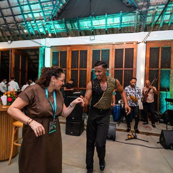 Ein Mann und eine Frau tanzen in einem hell beleuchteten Raum, mit einer Band im Hintergrund. Die Band spielt Gitarren und Schlagzeug. Die Frau trägt ein braunes Kleid, und der Mann trägt eine Paillettenweste und Hose.