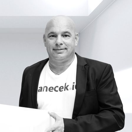 Zwei Männer stehen in einem Raum, einer hält eine große weiße Box. Einer trägt Anzug und Brille, der andere ein Shirt mit einem witzigen Zitat und einem 'anecek.io'-Logo. Sie schauen direkt in die Kamera.