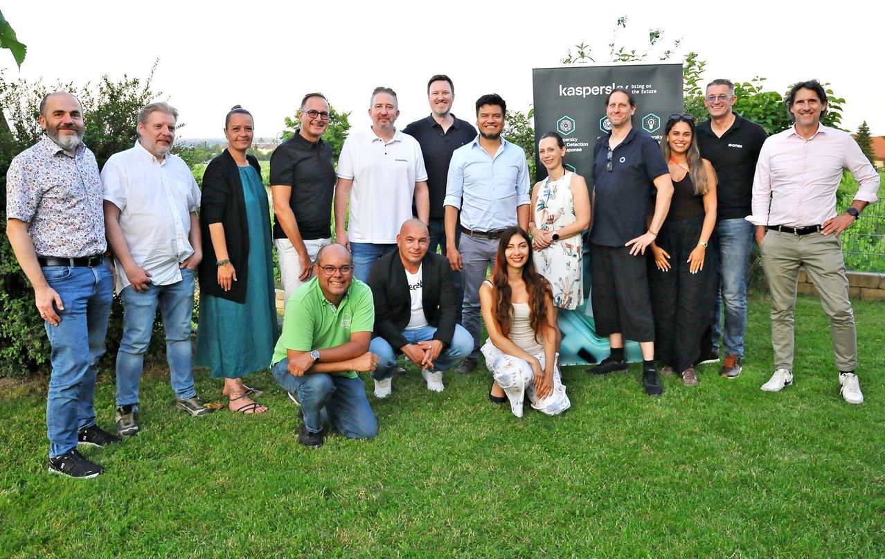 Gruppenfoto von verschiedenen Menschen, die auf Gras stehen und sitzen, einige mit Brille, vor einem Kaspersky-Banner.