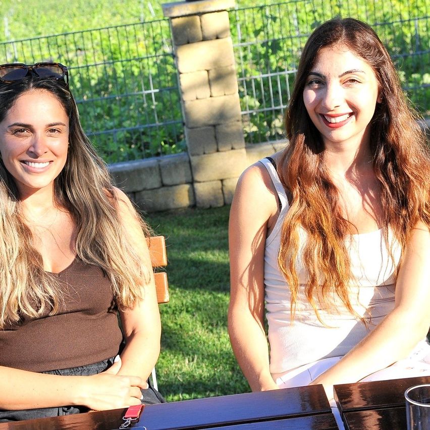 Zwei Frauen sitzen lächelnd an einem Tisch im Freien. Eine trägt eine Sonnenbrille auf dem Kopf und ein braunes Oberteil. Die andere trägt ein weißes Oberteil mit langen welligen Haaren. Ein Glas steht auf dem Tisch.