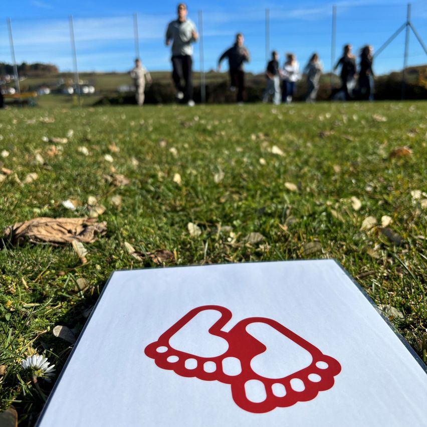 A group of people are running on a grassy field. In the foreground, there is a white paper with a red paw print design.