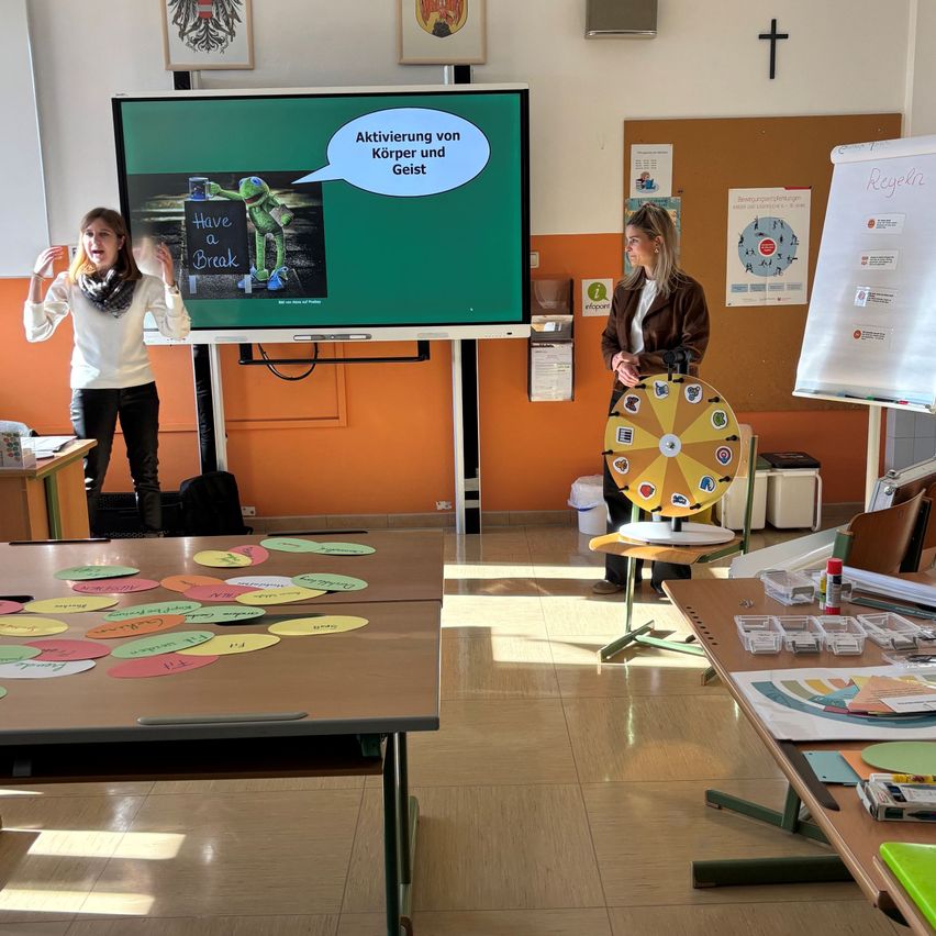 Two women in a classroom, one presents on a large screen while the other stands next to a colorful wheel and a whiteboard. Various papers are scattered on the tables.