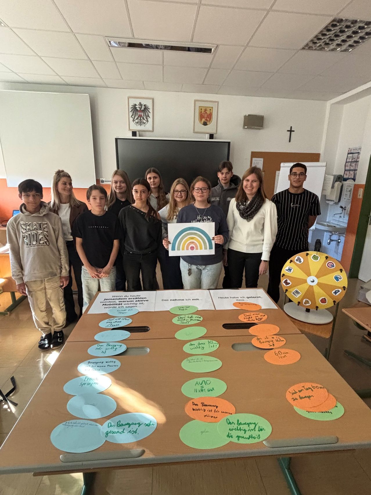 A group of students is posing in a classroom with a table displaying colorful circles, each labeled with activities like 'Walking' and 'Biking.' They are holding a colorful wheel and smiling.