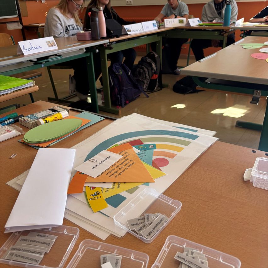 A classroom with several students seated at desks. On the desks, there are name tags, books, and backpacks. A colorful chart is on the desk in the foreground, and various papers are scattered.