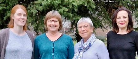 Vier Frauen stehen auf einem Rasen, lächelnd, mit einem Baum und einem Gebäude im Hintergrund.