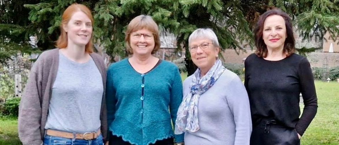 Vier Frauen posieren für ein Foto in einem Garten mit einem Baum im Hintergrund. Sie alle lächeln und scheinen für ein Bild zu posieren.