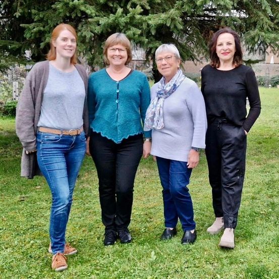 Vier Frauen posieren für ein Foto auf einer Rasenfläche mit Bäumen im Hintergrund. Sie tragen alle Freizeitkleidung.