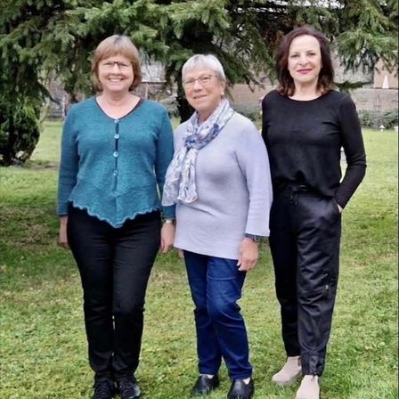 Drei Frauen stehen zusammen auf Gras in einem Park. Sie lächeln und tragen Freizeitkleidung. Eine trägt eine Brille.