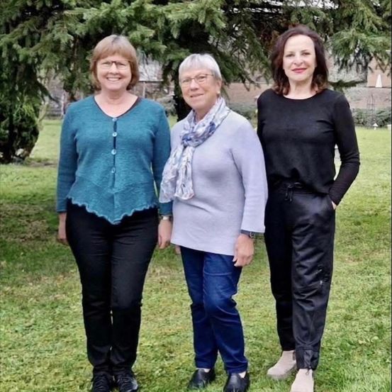 Drei Frauen stehen zusammen auf Gras in einem Park. Sie lächeln und tragen Freizeitkleidung. Eine trägt eine Brille.