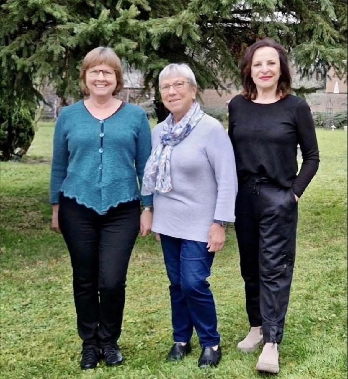 Drei Frauen stehen auf einem Rasen und posieren lächelnd für ein Foto. Die Frau links trägt ein blaues Oberteil und schwarze Hosen. Die Frau in der Mitte trägt ein graues Langarmshirt, Jeans und eine Krawatte. Die Frau rechts trägt ein schwarzes Langarmshirt und schwarze Hosen. Sie stehen vor einem Gebäude mit Bäumen.