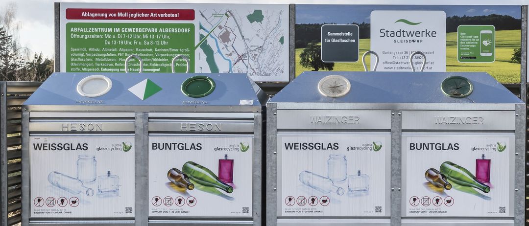 Zwei Glasrecyclingbehälter, einer für weißes und einer für farbiges Glas, stehen im Freien. Ein großer Plan und ein Schild, das Stadtwerke Gleisdorf bewirbt, sind im Hintergrund.