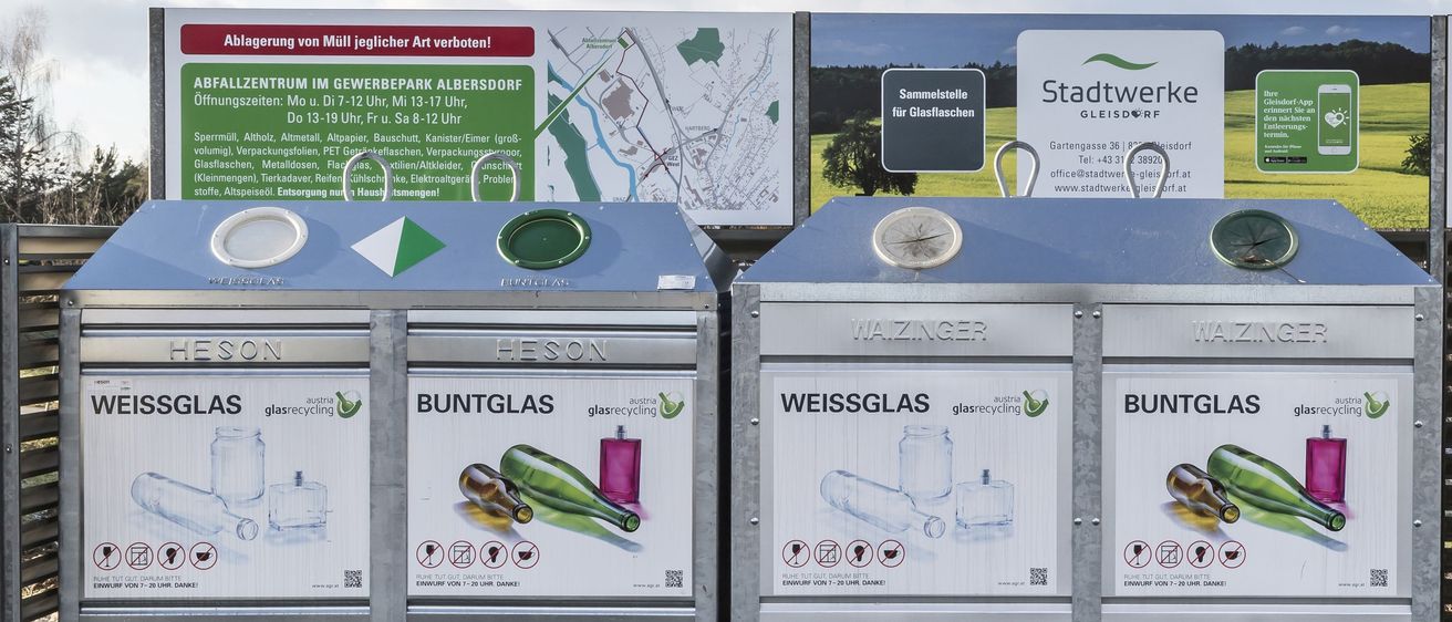 Zwei Glasrecyclingbehälter, einer für weißes und einer für farbiges Glas, stehen im Freien. Ein großer Plan und ein Schild, das Stadtwerke Gleisdorf bewirbt, sind im Hintergrund.