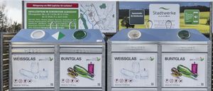 Zwei Glasrecyclingbehälter, einer für weißes und einer für farbiges Glas, stehen im Freien. Ein großer Plan und ein Schild, das Stadtwerke Gleisdorf bewirbt, sind im Hintergrund.