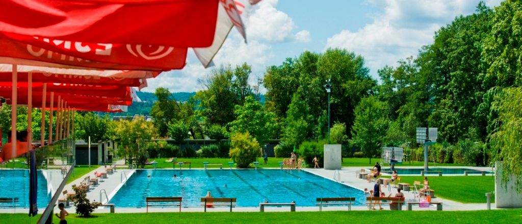 Ein Freibad umgeben von saftigem grünem Gras und Bäumen, mit Menschen, die schwimmen und sich auf Bänken unter roten Sonnenschirmen entspannen.