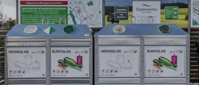 Zwei Recyclingbehälter stehen nebeneinander. Der linke Behälter hat ein Etikett für weißes Glas und der rechte für farbiges Glas. Beide sind mit Weissglas und Buntglas beschriftet. Dahinter befindet sich ein großes Banner mit verschiedenen Texten und Logos.