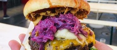 Eine Nahaufnahme eines Burgers mit Rinderpatty, Käse und roter Kohl wird in der Hand gehalten, mit bunten Zelten im Hintergrund.